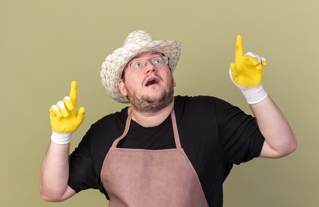 Asustado mirando hacia arriba joven jardinero vistiendo guantes y sombrero de jardinería apunta hacia arriba aislado en la pared verde oliva