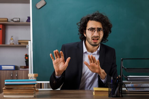 Asustado mostrando gesto de parada maestro con gafas sentado a la mesa con herramientas escolares en el aula