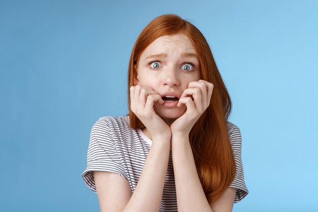 Asustado sin palabras aturdido asustado niña pelirroja temblor miedo ojos abiertos aterrorizado morderse las uñas fruncir el ceño temblando ansiosamente de pie fondo azul jadeando sorprendido