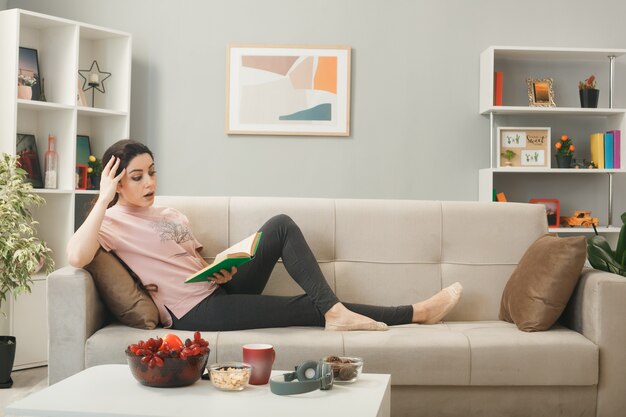 Asustado poner la mano en la cabeza joven tumbado en el sofá detrás de la mesa de café libro de lectura en la sala de estar