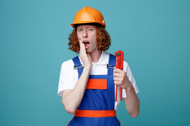 Asustado poniendo la mano en la boca joven constructor hombre en uniforme con llave de gas aislado sobre fondo azul.