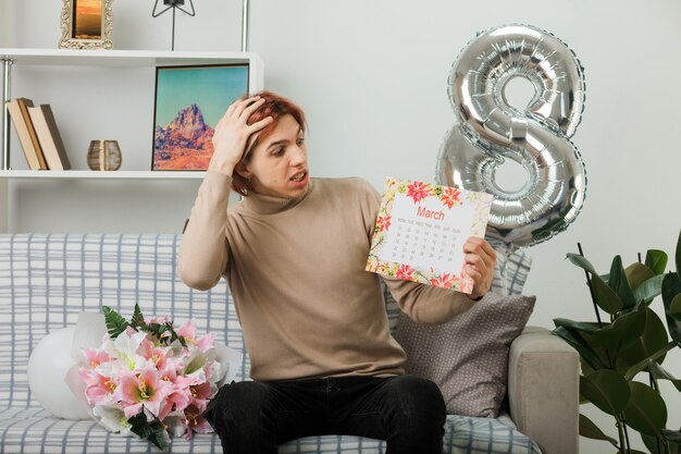 Asustado poniendo la mano en la cabeza chico guapo en el día de la mujer feliz sosteniendo y mirando el calendario sentado en el sofá en la sala de estar