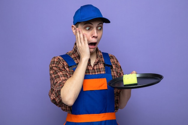 Foto gratuita asustado poniendo la mano en la mejilla joven chico de limpieza con uniforme y gorra sosteniendo y mirando la esponja en la bandeja