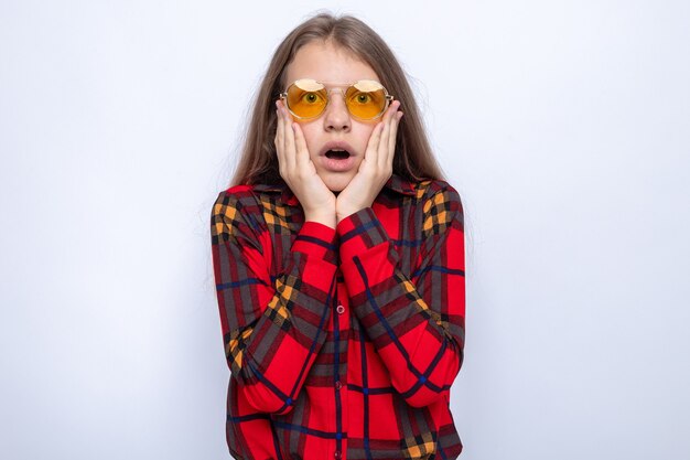 Asustado poniendo las manos en las mejillas hermosa niña vestida con camisa roja y gafas