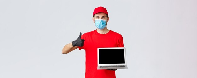 Atención al cliente covid19 paquetes de entrega concepto de procesamiento de pedidos en línea Mensajero alegre con mascarilla médica uniforme roja y guantes recomendar página web mostrar pantalla de computadora portátil y pulgar hacia arriba