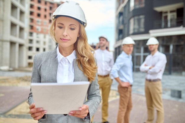 Atención. La empresaria con largo cabello rubio en casco de seguridad mirando de cerca el documento de pie en el sitio de construcción y el grupo de trabajo detrás