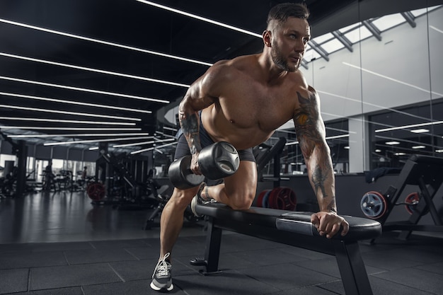 Atentado. Joven atleta caucásico muscular practicando en el gimnasio con las pesas. Modelo masculino haciendo ejercicios de fuerza, entrenando la parte superior del cuerpo. Bienestar, estilo de vida saludable, concepto de culturismo.