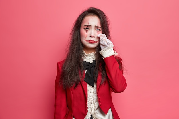 Aterradora mujer morena abatida con maquillaje de halloween enjuga lágrimas tiene expresión sombría El arte de la cara ensangrentada usa chaqueta roja y guantes de encaje Lente blanca en poses de ojos en el interior contra la pared rosada