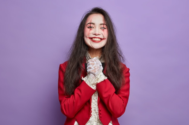 La aterradora niña de Halloween con maquillaje espeluznante sonríe con alegría anticipa la fiesta de disfraces mantiene las manos juntas aisladas en la pared púrpura Arte de cara ensangrentada