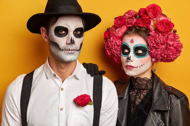 La aterradora niña zombie se inclina sobre el hombro del hombre, mira con atención, el hombre serio viste sombrero negro, camisa blanca con tirantes, se prepara para la celebración de Halloween.