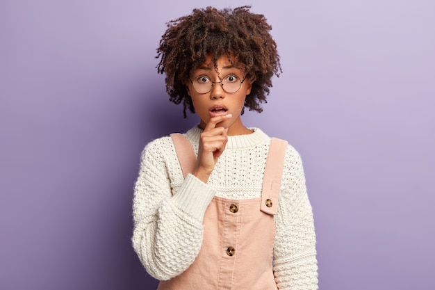 Aterrorizada joven de piel oscura con peinado afro, ha contenido el aliento, se ve sorprendentemente, escucha noticias impactantes