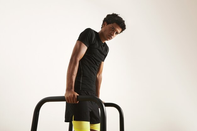 Atleta afroamericano musculoso joven serio en camiseta técnica negra y pantalones cortos negros y amarillos haciendo inmersiones en barras cortas aisladas en blanco.