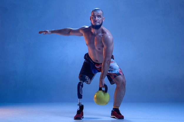 Atleta con discapacidad o amputado aislado en pared azul. Deportista masculino profesional con entrenamiento de prótesis de pierna con pesas en neón. Deporte para discapacitados y superación, concepto de bienestar.