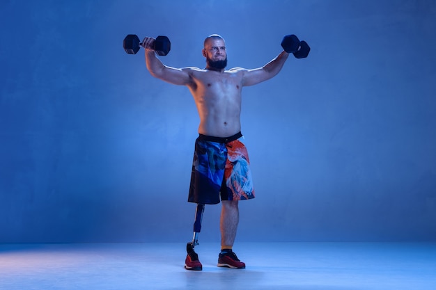 Foto gratuita atleta con discapacidad o amputado aislado en pared azul. deportista masculino profesional con entrenamiento de prótesis de pierna con pesas en neón. deporte para discapacitados y superación, concepto de bienestar.