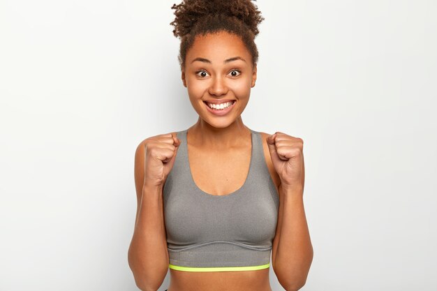 La atleta femenina aprieta los puños con alegría, celebra la victoria del maratón, sonríe ampliamente, vestida con un top gris, sonríe ampliamente, disfruta del triunfo, posa en el interior contra la pared blanca