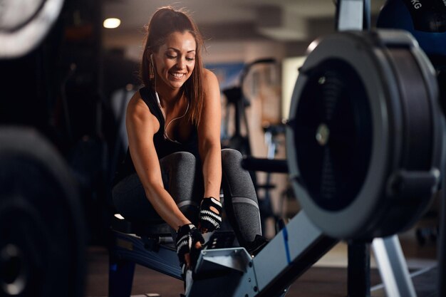 Atleta femenina feliz ajustando la máquina de remo y escuchando música en los auriculares durante el entrenamiento deportivo en un gimnasio