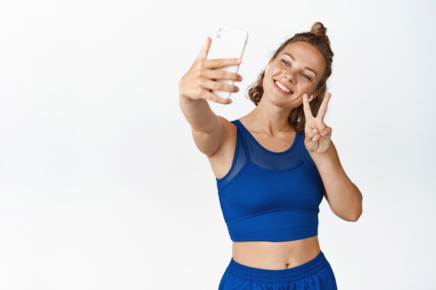 Atleta femenina feliz tomando selfie en teléfono móvil, fotografiando en auriculares y ropa activa, fondo blanco