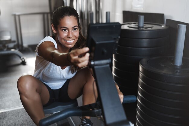 Atleta femenina joven motivada, sonriendo en el gimnasio, usando equipo de prensa de piernas