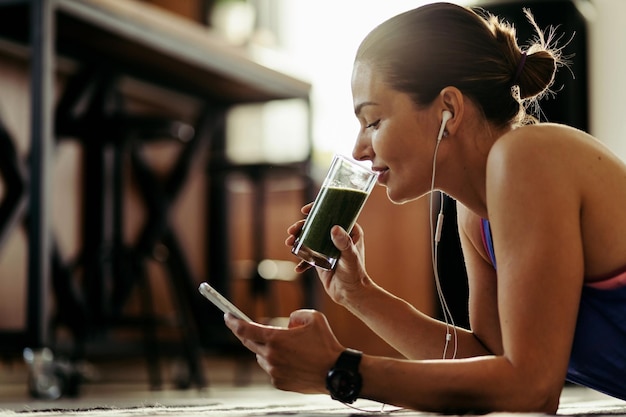 Atleta femenina sonriente usando un teléfono inteligente y bebiendo un batido saludable mientras se acuesta en el suelo en casa