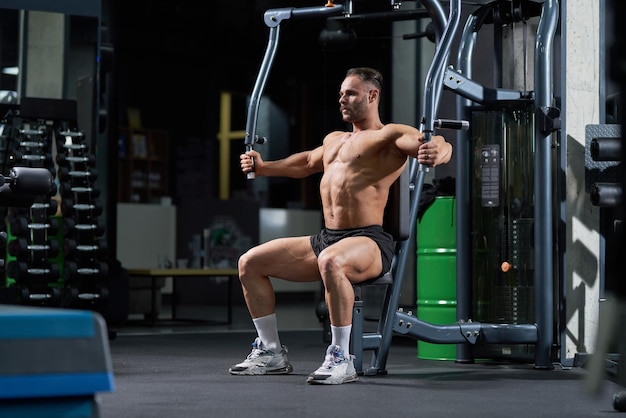 Foto gratuita atleta fuerte sin camisa sentado mientras entrena en una máquina crossover en el gimnasio vista lateral de caucásico