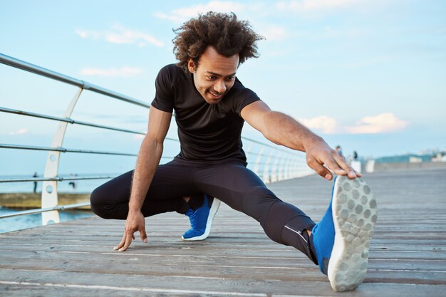 Atleta hombre de piel oscura en ropa deportiva negra y zapatillas azules estirando sus piernas con ejercicio de estiramiento de isquiotibiales en el muelle. Calentamiento del corredor masculino joven afroamericano