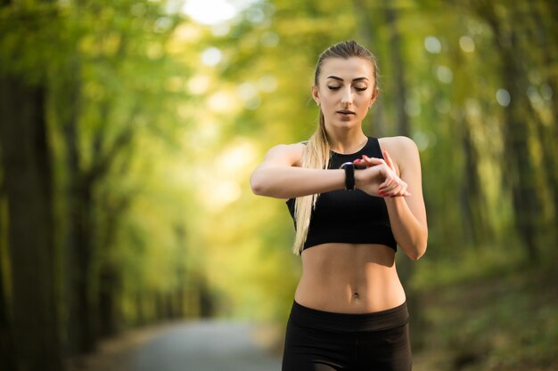 Atleta joven atractiva practicando fitness al aire libre y configurando su monitor de cardio smartwatch antes de correr en el parque