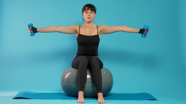 Atleta levantando pesas y sentado en una pelota tonificante de fitness en el estudio, entrenando músculos de brazos con pesas y equipos deportivos. Mujer activa haciendo ejercicio y estirándose con actividad de entrenamiento.