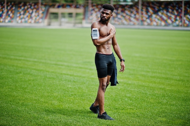 Atleta masculino afroamericano sexy deporte torso desnudo hombre con estuche de brazo deportivo para teléfono móvil posado en la hierba verde del estadio de fútbol