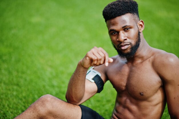 Atleta masculino afroamericano sexy deporte torso desnudo hombre con estuche de brazo deportivo para teléfono móvil posado en la hierba verde del estadio de fútbol