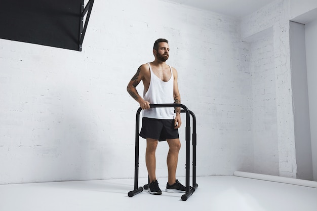 Atleta masculino barbudo tatuado brutal en camiseta sin mangas blanca en blanco de pie junto a barras paralelas y barra de tiro mirando de lado, listo para entrenar, mirando de lado