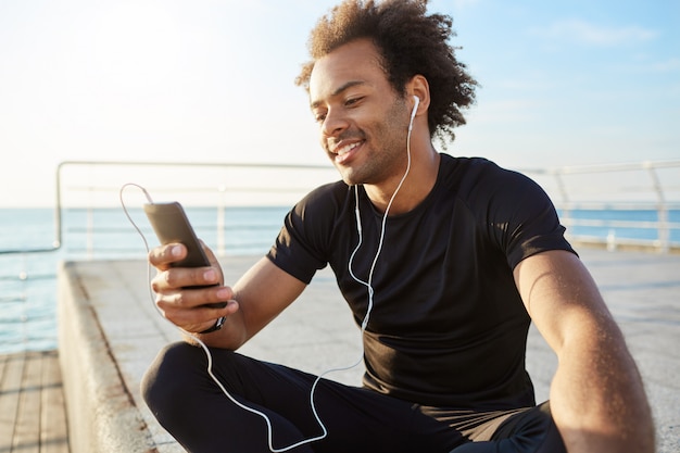 Atleta masculino elegante de piel oscura con peinado afro usando teléfono móvil, sonriendo, eligiendo la mejor canción para el entrenamiento