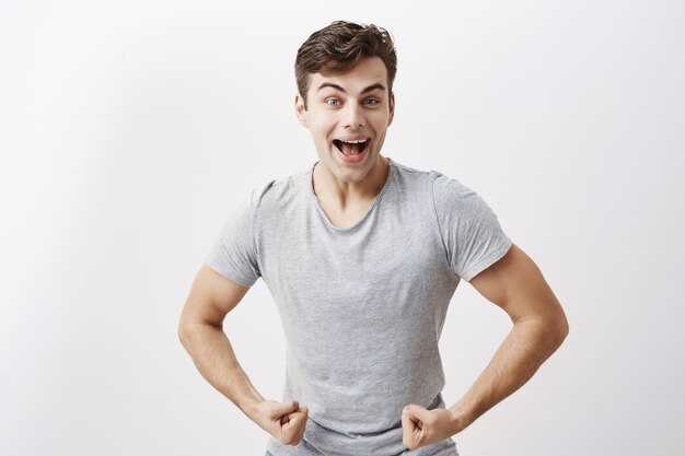 Atleta masculino joven emocional con cuerpo musculoso, ampliamente sonriendo, demostrando lo fuerte que es, se jacta de sí mismo. Chico positivo muestra sus músculos y fuerza, posando.