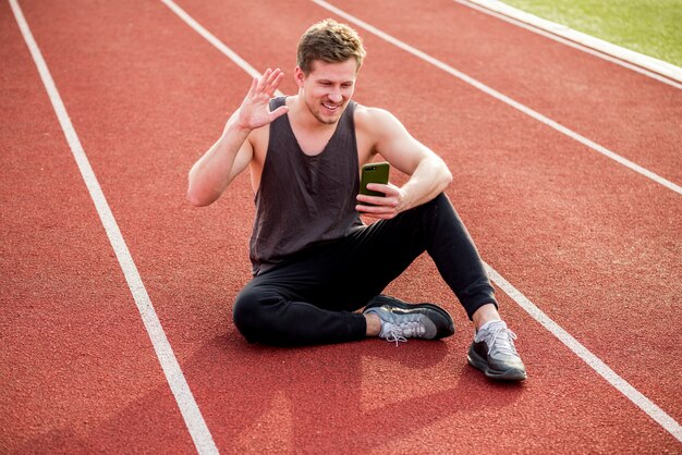 Atleta masculino joven sonriente que se sienta en pista roja que agita sus manos mientras que hace la videollamada