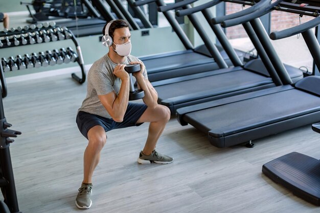 Atleta masculino con mascarilla usando el peso de la mano mientras hace sentadillas en el club de salud