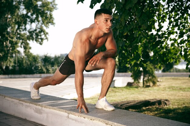 Un atleta masculino musculoso haciendo ejercicio en el parque. Gimnasia, entrenamiento, entrenamiento físico, flexibilidad. Ciudad de verano en un día soleado en el campo de fondo. Estilo de vida activo y saludable, juventud, culturismo.