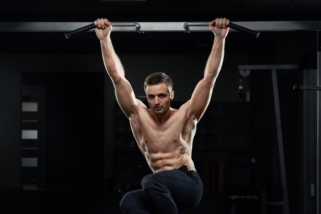 Atleta masculino musculoso tirando hacia arriba en la barra horizontal en el gimnasio oscuro