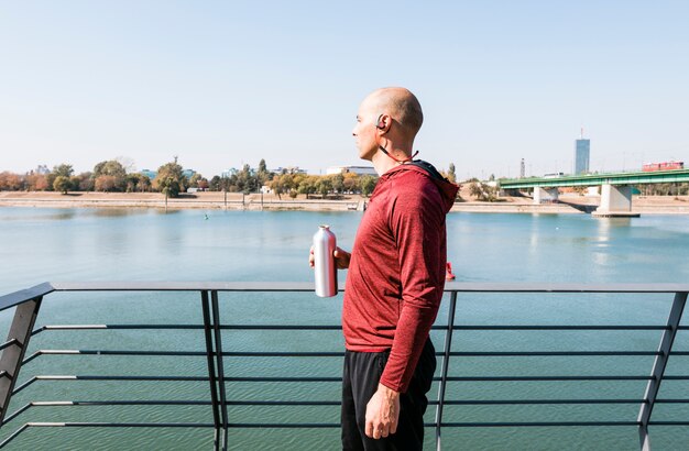 Un atleta masculino que lleva bluetooth inalámbrico en su oreja sosteniendo una botella de agua en la mano mirando a la vista