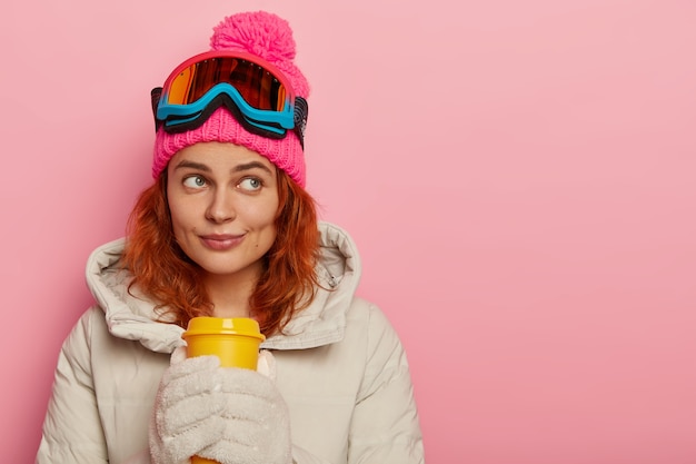 Atleta mujer pensativa, viste ropa abrigada, bebe café para llevar, concentrado, modelos contra la pared rosa del estudio