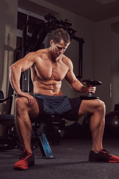 Un atleta musculoso sin camisa haciendo ejercicio con pesas mientras se sienta en un banco en el gimnasio.