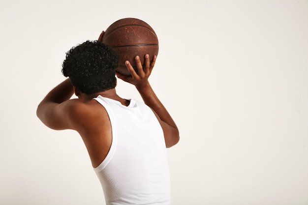 Atleta musculoso de piel oscura con un afro y una diadema con camisa blanca sin mangas lanzando una vieja pelota de baloncesto de cuero marrón sobre blanco
