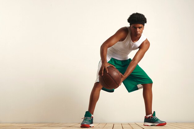 Atleta negro serio en verde y blanco con una pelota de baloncesto vintage sostenida contra su rodilla