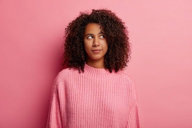 Atractiva adolescente con cabello afro mira pensativamente en la esquina superior derecha, tiene expresión pensativa, vestida con un suéter rosa, plantea en el interior, dudas sobre algo.