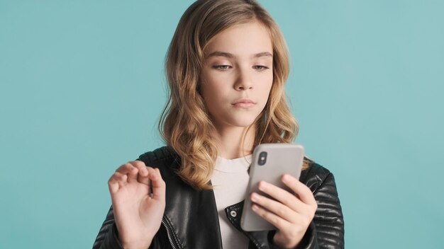 Atractiva adolescente rubia enviando un mensaje a un amigo usando un teléfono inteligente sobre fondo azul