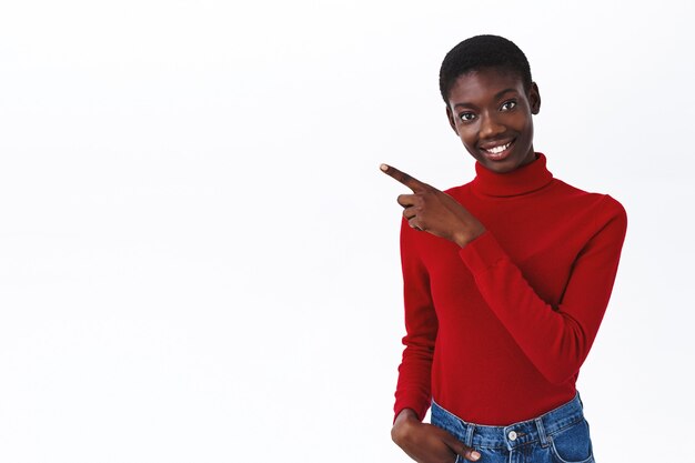 Atractiva y amigable modelo de mujer afroamericana en cuello alto rojo da consejos, apuntando con el dedo a la izquierda en el espacio en blanco en blanco