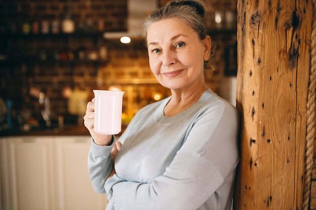 Atractiva anciana alegre posando en el interior disfrutando de un café recién hecho caliente de la taza por la mañana.