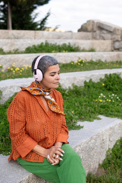 Atractiva anciana escuchando música en el parque. Modelo femenino con cabello gris corto en ropa brillante y grandes auriculares. Ocio, actividad, concepto de tecnología.