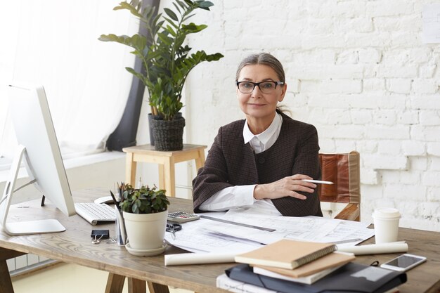 Atractiva arquitecta madura con gafas disfrutando del proceso de trabajo en una oficina espaciosa y luminosa, sentada frente a una computadora genérica, sosteniendo un lápiz, examinando dibujos y especificaciones en el escritorio