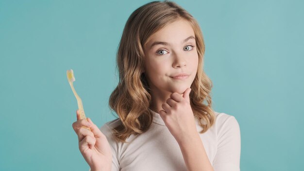 Atractiva chica adolescente de pelo ondulado rubio sosteniendo un cepillo de dientes mirando pensativo sobre fondo azul. expresión de pensamiento