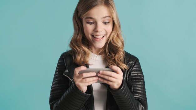 Atractiva chica adolescente rubia emocionalmente jugando juegos en el teléfono inteligente que se ve feliz aislada en el fondo azul