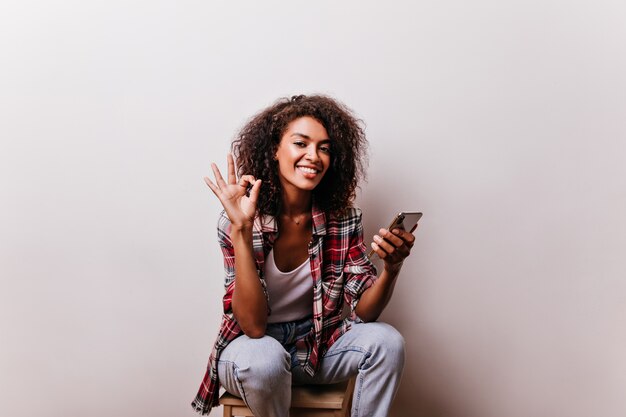 Atractiva chica africana posando con signo bien. Señora negra rizada con sonrisa romántica con smartphone.